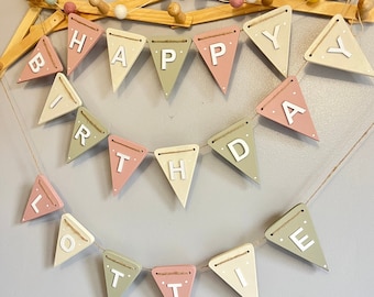 Op de afbeelding: Een houten banner met de woorden "Happy Birthday Lottie" in witte letters op een roze, groene en crème achtergrond. De banner is aan een touwtje geregen en heeft kleine witte stippen op elke vlag.
