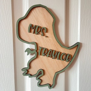 May include: A wooden dinosaur cutout with a green outline. The dinosaur is holding a sign that says "Mrs. Traylor".