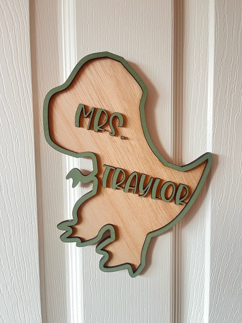 May include: A wooden dinosaur cutout with a green outline. The dinosaur is holding a sign that says "Mrs. Traylor".