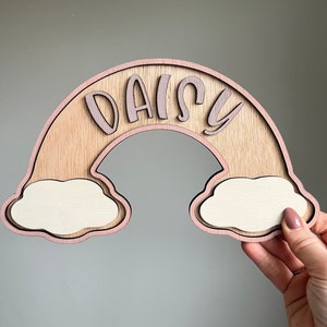 May include: A wooden rainbow with a pink outline and the name "DAISY" cut out in the centre. The rainbow has two white clouds attached to the bottom.