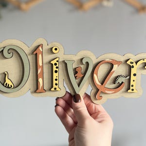 K&ouml;nnte beinhalten: Ein h&ouml;lzernes Namensschild mit dem Namen "Oliver", der in bunten Buchstaben mit Tierillustrationen ausgeschrieben ist. Die Buchstaben sind in Gr&uuml;n-, Gelb-, Orange- und Braunt&ouml;nen bemalt.