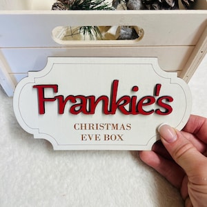 May include: A decorative wooden plaque with a cream-coloured finish and a scalloped edge. The text "Frankie's" is displayed in raised, bold red lettering, while the words "CHRISTMAS EVE BOX" are printed in brown below the name.