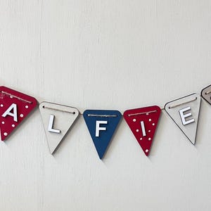Może przedstawiać: Drewniany baner z napisem "ALFIE" w postaci pojedynczych tr&oacute;jkątnych flag. Flagi są w r&oacute;żnych kolorach, w tym czerwonym z białymi kropkami, beżowym i niebieskim. Każda flaga ma literę lub element dekoracyjny, połączone białym sznurkiem.