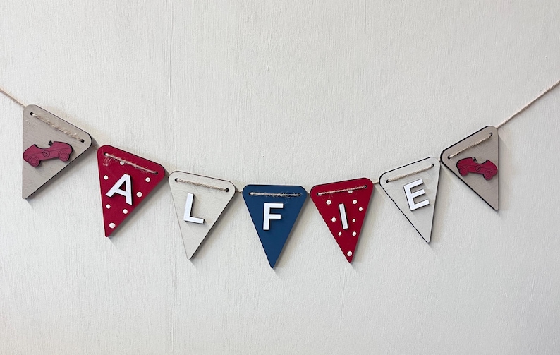 Może przedstawiać: Drewniany baner z napisem "ALFIE" w postaci pojedynczych tr&oacute;jkątnych flag. Flagi są w r&oacute;żnych kolorach, w tym czerwonym z białymi kropkami, beżowym i niebieskim. Każda flaga ma literę lub element dekoracyjny, połączone białym sznurkiem.