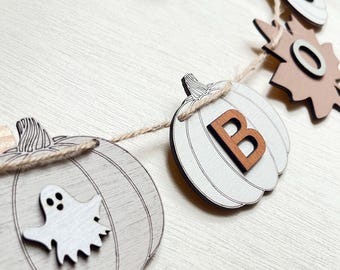 May include: A decorative garland featuring light gray wooden pumpkin cutouts strung on twine. One pumpkin displays a white ghost figure, while another features a dark brown letter B. Additional shapes include a brown leaf and a white pumpkin.