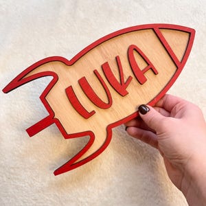 May include: A wooden rocket ship with a red outline and the name "Luka" written in red letters.
