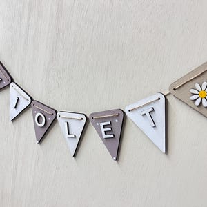 May include: A wooden banner spelling "VIOLET" in white capital letters on grey and white pennants. The banner is strung on twine and features daisy accents on the end pennants. The background is a light wood grain.