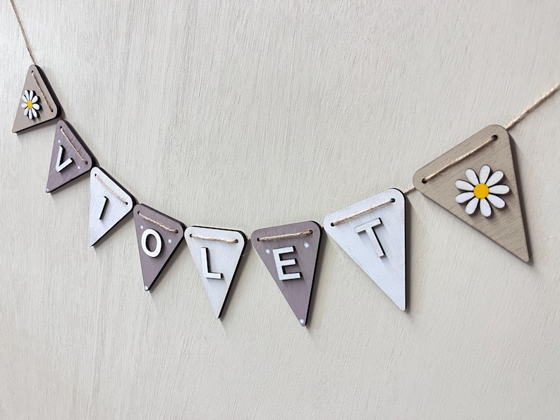 May include: A wooden banner spelling "VIOLET" in white capital letters on grey and white pennants. The banner is strung on twine and features daisy accents on the end pennants. The background is a light wood grain.