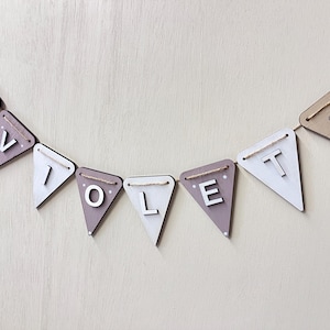 May include: A wooden banner spelling "VIOLET" with individual triangle flags. The flags alternate between white and a muted purple-grey colour. Each flag is strung on a natural twine. Some flags feature a daisy.