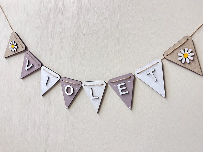 May include: A wooden banner spelling "VIOLET" with individual triangle flags. The flags alternate between white and a muted purple-grey colour. Each flag is strung on a natural twine. Some flags feature a daisy.