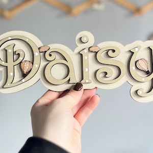 K&ouml;nnte beinhalten: Ein Holzschild mit dem Namen "Daisy", der in Kursivschrift geschrieben ist. Die Buchstaben sind in einem hellen Braunton lackiert und haben wei&szlig;e Punkte an den R&auml;ndern. An den Buchstaben sind kleine, braune Hei&szlig;luftballons befestigt.