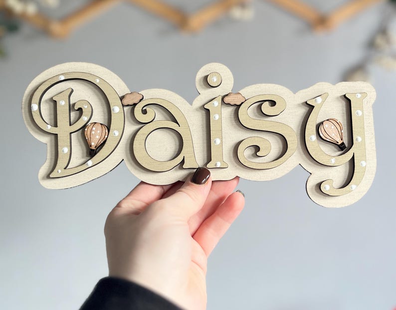 K&ouml;nnte beinhalten: Ein Holzschild mit dem Namen "Daisy", der in Kursivschrift geschrieben ist. Die Buchstaben sind in einem hellen Braunton lackiert und haben wei&szlig;e Punkte an den R&auml;ndern. An den Buchstaben sind kleine, braune Hei&szlig;luftballons befestigt.