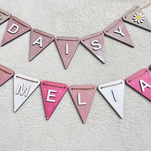 May include: Wooden bunting spelling "DAISY" and "AMELIA" in white capital letters on pink, white, and grey pennant flags. Each end flag features a daisy. The flags are strung on twine, ideal for a nursery or party.