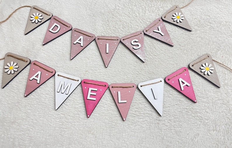May include: Wooden bunting spelling "DAISY" and "AMELIA" in white capital letters on pink, white, and grey pennant flags. Each end flag features a daisy. The flags are strung on twine, ideal for a nursery or party.