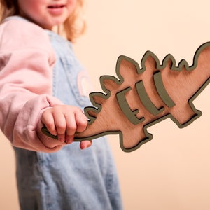 May include: A child holds a wooden dinosaur cutout painted in a light green color. The dinosaur has a textured wood grain and is shaped like a stegosaurus.