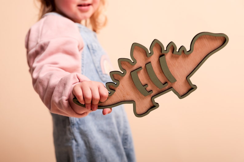 May include: A child holds a wooden dinosaur cutout painted in a light green color. The dinosaur has a textured wood grain and is shaped like a stegosaurus.