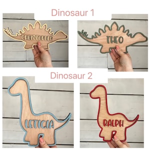 May include: Four wooden dinosaur-shaped plaques with names written on them. The dinosaurs are in different colors, including green, blue, red, and yellow. The names are Christopher, Theo, Leticia, and Ralph.