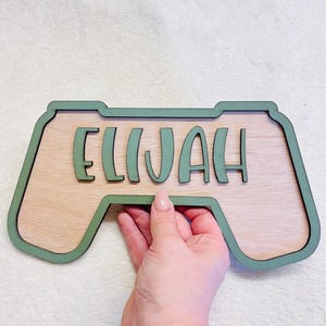May include: A wooden wall sign shaped like a video game controller with a light wood grain centre and a sage green border. The name ELIJAH is cut out in sage green letters and mounted on the centre of the controller-shaped wooden plaque.