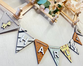 Op de afbeelding: Een decoratieve slinger met driehoekige houten vlaggetjes met op dieren geïnspireerde patronen en de naam HARLEY in witte, verhoogde letters. De vlaggetjes zijn verbonden met touw en hangen aan een lichtgekleurd houten wandrek.