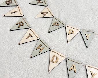 May include: A wooden banner spelling "BIRTHDAY" with individual triangle flags. The flags alternate between light green and natural wood tones, with gold-colored letters. The banner is strung on a natural twine.