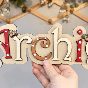 K&ouml;nnte beinhalten: Ein h&ouml;lzernes Namensschild mit dem Namen "Archie" in wei&szlig;en Buchstaben mit roten und wei&szlig;en Punkten. Die Buchstaben sind &uuml;bereinander gelegt und haben einen 3D-Effekt. Auf dem Schild befinden sich zwei kleine Spielzeugtraktoren.