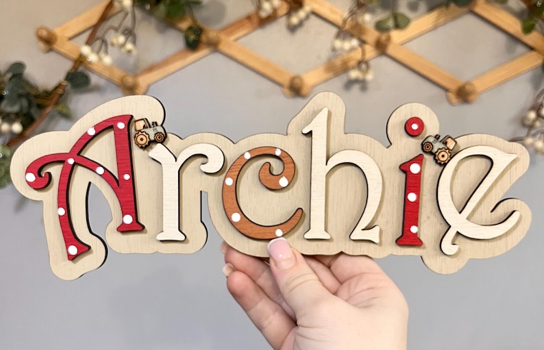 K&ouml;nnte beinhalten: Ein h&ouml;lzernes Namensschild mit dem Namen "Archie" in wei&szlig;en Buchstaben mit roten und wei&szlig;en Punkten. Die Buchstaben sind &uuml;bereinander gelegt und haben einen 3D-Effekt. Auf dem Schild befinden sich zwei kleine Spielzeugtraktoren.
