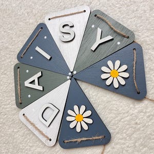 May include: Wooden banner spelling "DAISY" in white letters on triangular flags. The flags are painted in shades of blue, green, and white, with daisy accents and twine for hanging.