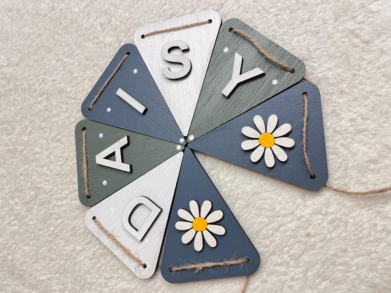 May include: Wooden banner spelling "DAISY" in white letters on triangular flags. The flags are painted in shades of blue, green, and white, with daisy accents and twine for hanging.