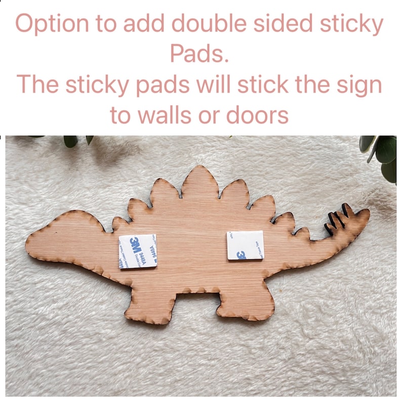 May include: A wooden dinosaur cutout with two 3M adhesive pads attached to the back. The text "Option to add double sided sticky Pads. The sticky pads will stick the sign to walls or doors" is written above the dinosaur.