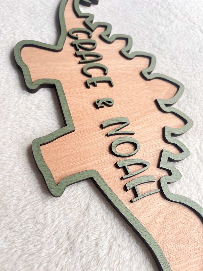 May include: A wooden dinosaur-shaped sign with a green outline. The sign says "GRACE & NOAH" in a green cursive font.
