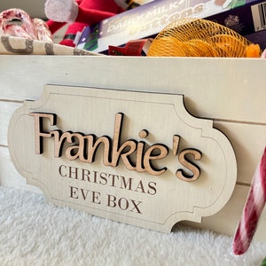 May include: A light-coloured wooden sign with the text "Frankie's Christmas Eve Box". The sign has a decorative shape and raised lettering. The box is filled with Christmas treats and an elf.
