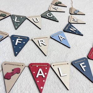 Może przedstawiać: Drewniany baner z tr&oacute;jkątnymi flagami, kt&oacute;re składają się na napis "LUCA FRALF". Każda flaga jest w innym kolorze, w tym beżowym, niebieskim, czerwonym i zielonym, i ma wz&oacute;r samochodu lub literę. Flagi połączone są naturalnym sznurkiem.