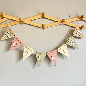 May include: A pastel-coloured wooden banner spelling "CALLIE" in white letters, strung with twine. The pennants are painted in pink, beige, and green. Some pennants have white polka dots. Two pennants feature a daisy. The banner hangs on a wooden wall rack.