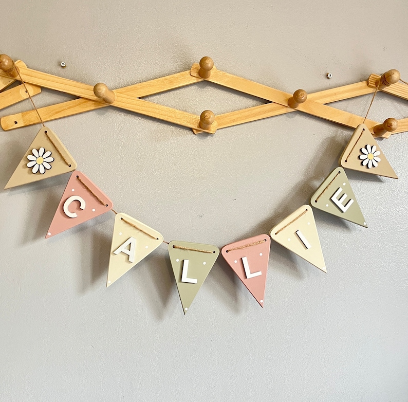 May include: A pastel-coloured wooden banner spelling "CALLIE" in white letters, strung with twine. The pennants are painted in pink, beige, and green. Some pennants have white polka dots. Two pennants feature a daisy. The banner hangs on a wooden wall rack.