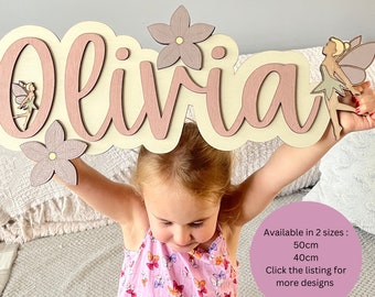 Könnte beinhalten: Ein hölzernes Namensschild mit dem Namen "Olivia" in rosa Schrift. Das Schild ist mit Blumen und Feen verziert. Erhältlich in zwei Größen: 50 cm und 40 cm. Klicken Sie auf die Liste für weitere Designs.