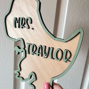 May include: A wooden dinosaur cutout with a green outline. The dinosaur is holding a sign that says "Mrs. Traylor".