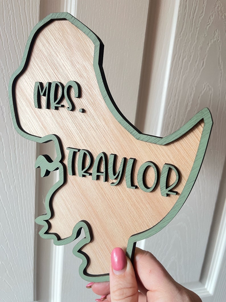 May include: A wooden dinosaur cutout with a green outline. The dinosaur is holding a sign that says "Mrs. Traylor".