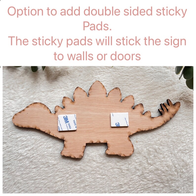 May include: A wooden dinosaur cutout with two 3M adhesive pads attached to the back. The text "Option to add double sided sticky Pads. The sticky pads will stick the sign to walls or doors" is written above the dinosaur.