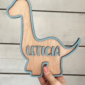 May include: A wooden dinosaur cutout painted light blue with the name "LETICIA" cut out in the center.