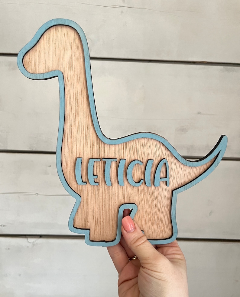 May include: A wooden dinosaur cutout painted light blue with the name "LETICIA" cut out in the center.