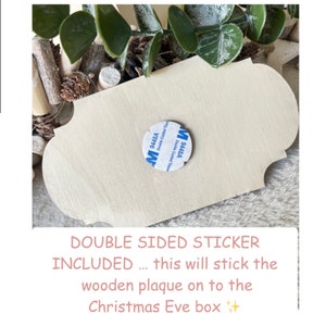 May include: A close-up of a wooden plaque with a double-sided sticker attached to the back. The sticker is labeled "9448A Double Coated Tissue". The text "DOUBLE SIDED STICKER INCLUDED... this will stick the wooden plaque on to the Christmas Eve box" is written below the image.