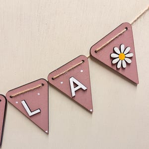 May include: A pink wooden banner with white letters spelling "LA" and a daisy. The banner is strung with twine. The daisy has white petals and a yellow centre. The banner is a decorative item.