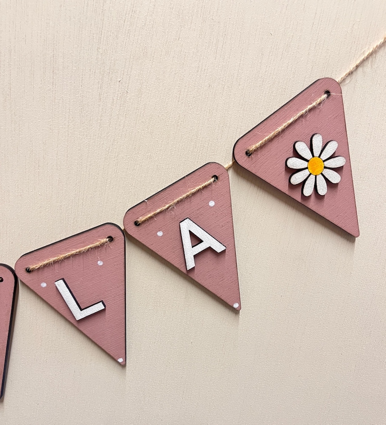 May include: A pink wooden banner with white letters spelling "LA" and a daisy. The banner is strung with twine. The daisy has white petals and a yellow centre. The banner is a decorative item.