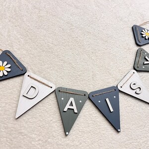 May include: A wooden banner spelling "DAISY" with individual triangle flags. The flags are painted in shades of white, grey, and navy blue. Some flags feature a daisy flower. The banner is strung with twine.