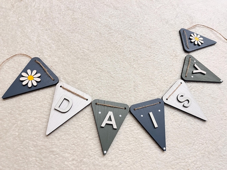 May include: A wooden banner spelling "DAISY" with individual triangle flags. The flags are painted in shades of white, grey, and navy blue. Some flags feature a daisy flower. The banner is strung with twine.