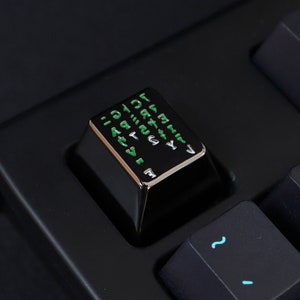 Könnte beinhalten: Eine schwarze mechanische Tastatur-Tastenkappe mit silberner Oberfläche und grüner Beschriftung. Die Tastenkappe hat eine Vielzahl von Zeichen und Symbolen, die darauf gedruckt sind.