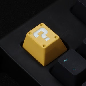 Peut inclure: Un capuchon de clavier jaune avec un point d'interrogation blanc dessus. Le capuchon est en forme de bloc d'interrogation du jeu vidéo Super Mario Bros.
