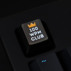 Könnte beinhalten: Eine schwarze mechanische Tastatur mit einer Tastenkappe, auf der "100 WPM CLUB" mit einem goldenen Kronensymbol steht. Die Tastenkappe ist silberfarben mit weißem Text.