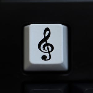 May include: A close-up shot of a white keyboard key featuring a black treble clef symbol. The key is set against a black background of other keys and the keyboard frame. The image is sharply focused.