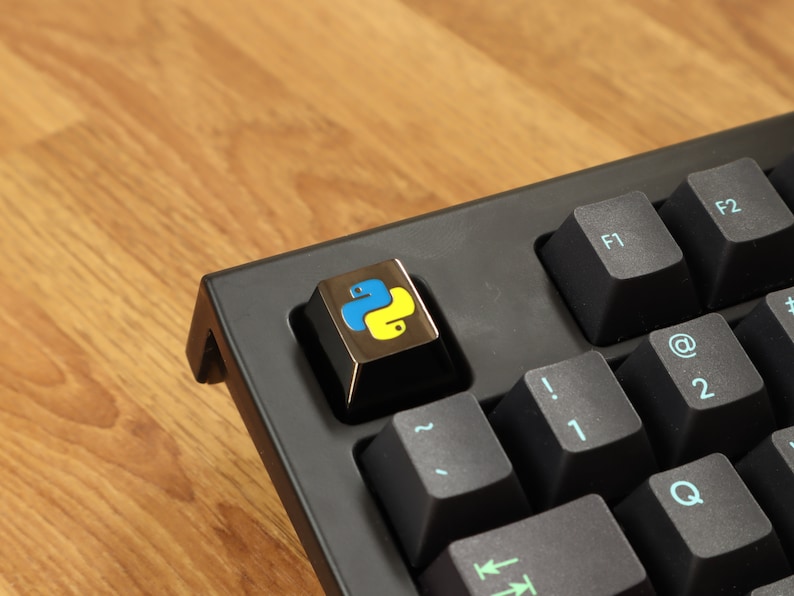 Pode incluir: Close-up de um teclado preto com uma tecla personalizada com o logotipo da linguagem de programa&ccedil;&atilde;o Python. A tecla &eacute; dourada com um emblema de cobra azul e amarelo. Outras teclas s&atilde;o pretas com letras azul claro.
